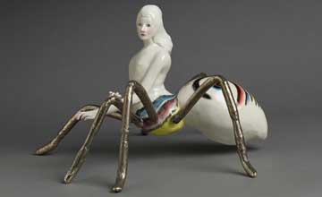 A ceramic spider with a woman's upper body standing on a grey backdrop.